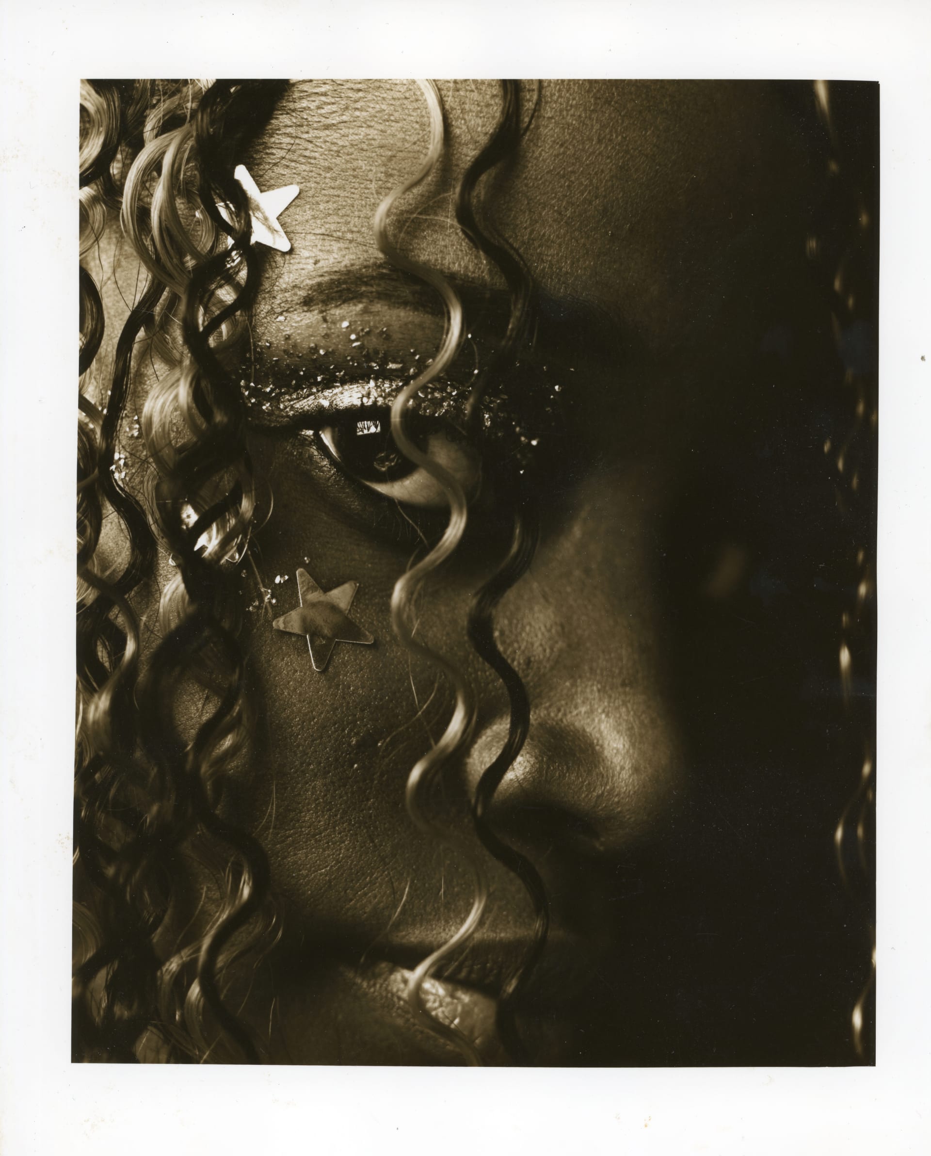 A close up tinted black and white photograph close up of the side of a young black woman's face. She is looking directly at the camera, she has coily hair covering her face and framing the image. She has small star shapes stuck to her face and glitter on her eyelids.
