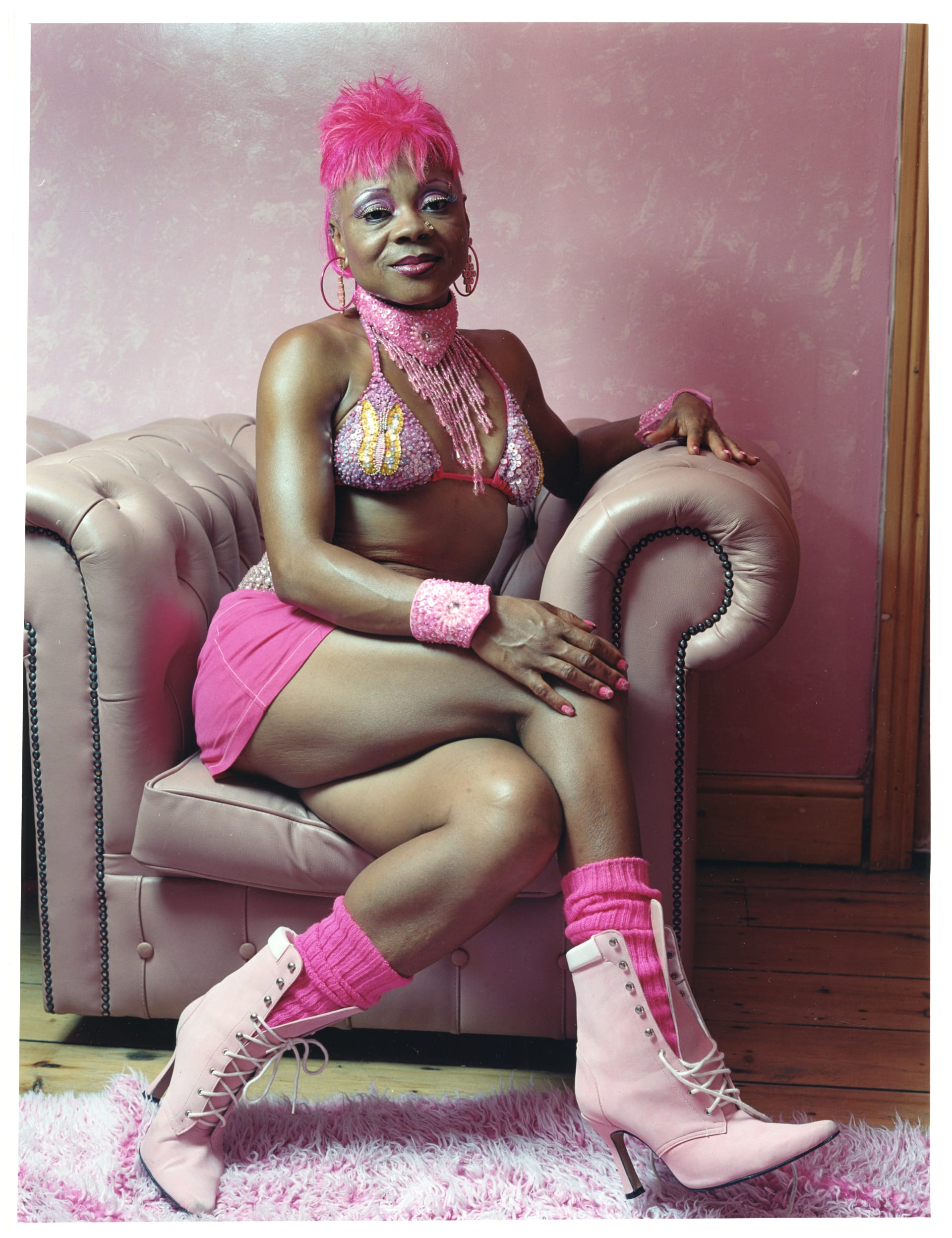 Colour photograph of a black woman wearing a bright pink wig, pink make-up, pink bikini top, pink necklace, earrings and bracelets, pink mini skirt, pink nails, pink socks, and high-heeled pink lace-up boots. She is relaxed and sitting cross-legged, looking directly at the camera sitting on a dusky pink leather armchair in a room with pink wallpaper, wooden floors and a pink fluffy rug in the foreground.
