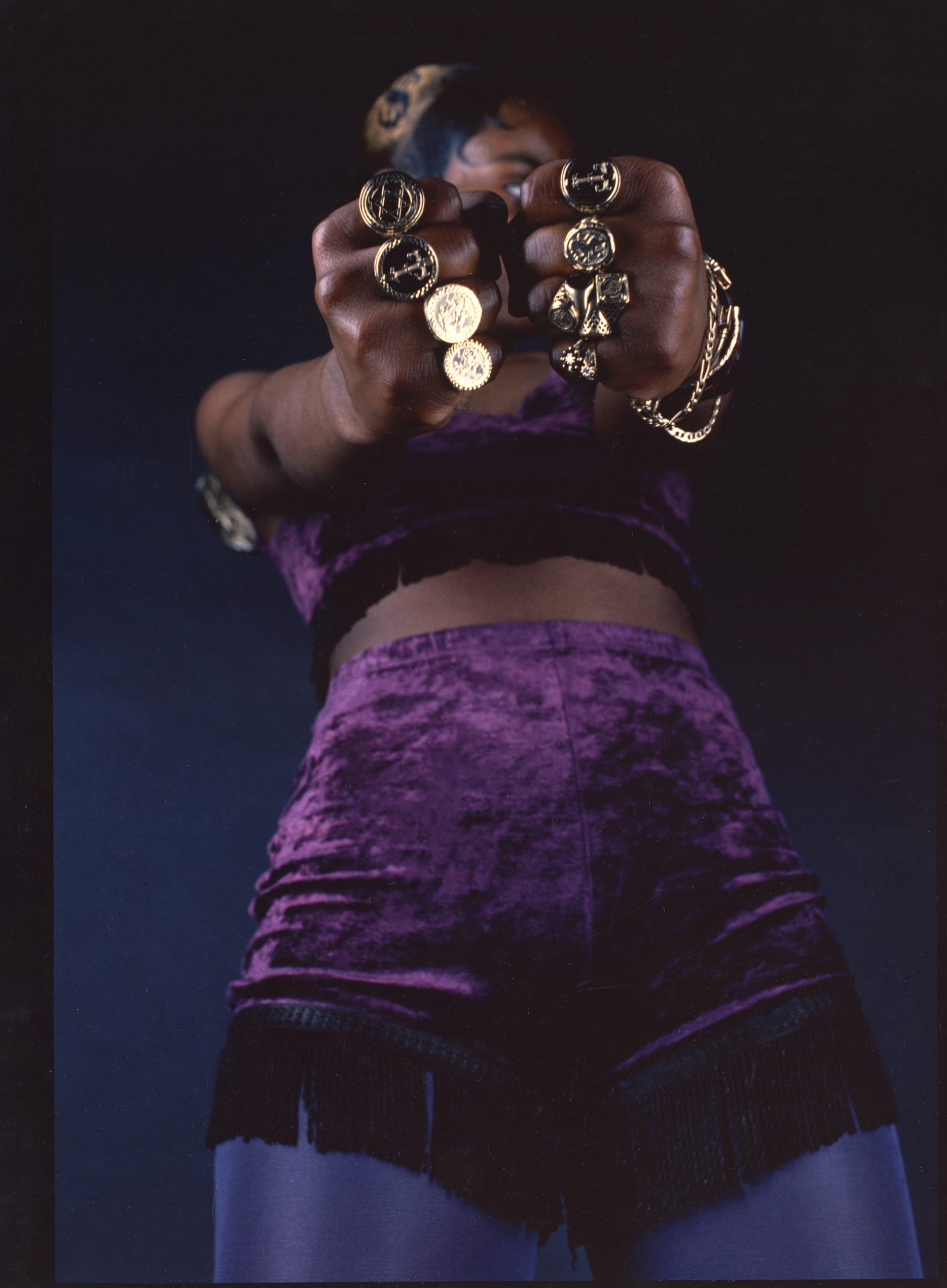 A colour photograph of a young black woman wearing purple hot pants and bikini top and purple tights against a dark background. Her hair is short, black, and gold and gelled tight to her head. The image is taken from below at a sharp angle. She is looking to her left and her face is obscured by her joined fists which are at the forefront of the image. On each finger is a significant-sized gold ring, many are round sovereign-shaped. She has gold bracelets on her left wrist.