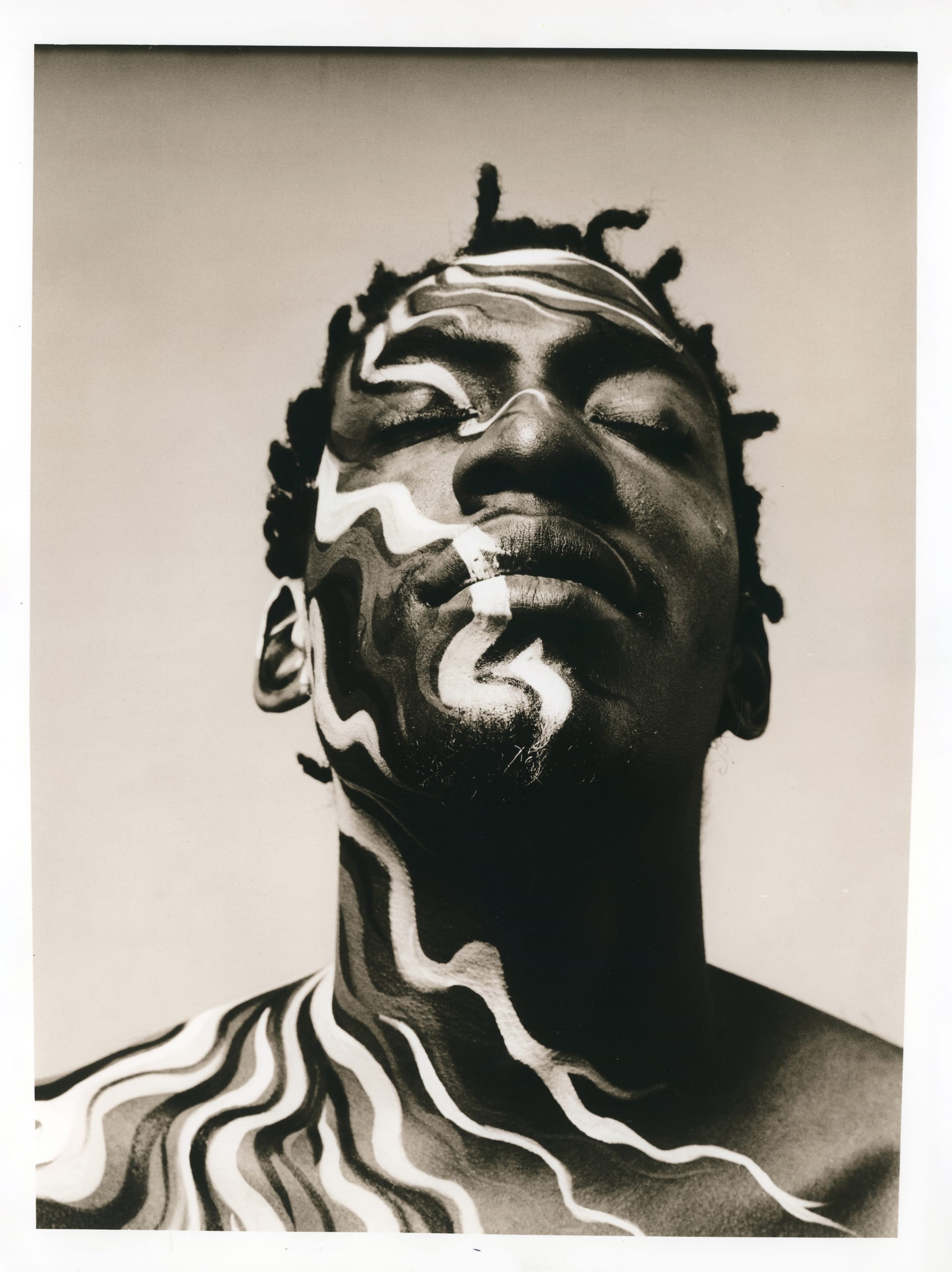 A black and white close-up portrait of rapper Roots Manuva. Head and shoulders with a detailed camouflage make-up. His head is tilted upwards and his eyes are closed.